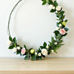 Minimalist floral hoop wreath with artificial roses, greenery, and delicate flowers, arranged asymmetrically on a thin wire frame, displayed on a wooden table against a white wall