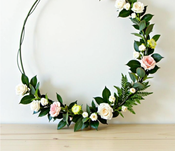 Minimalist floral hoop wreath with artificial roses, greenery, and delicate flowers, arranged asymmetrically on a thin wire frame, displayed on a wooden table against a white wall