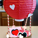 DIY Valentine's Day hot air balloon basket made from a wicker basket and red paper lantern, decorated with white hearts and filled with Valentine's gifts