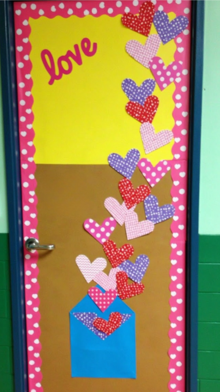 Classroom door decorated with a Valentine's Day theme, featuring a blue envelope at the bottom spilling out colorful patterned paper hearts, with 'love' written at the top.