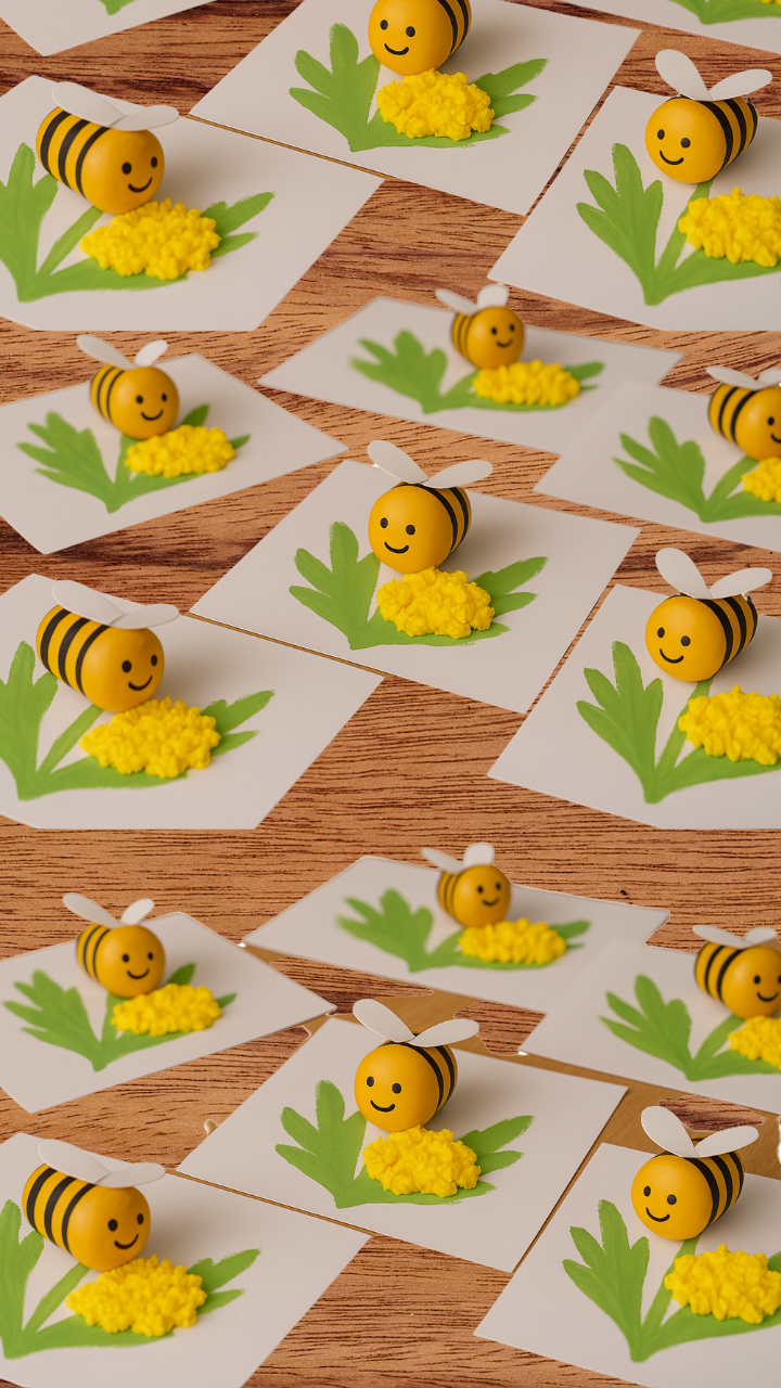Handmade bee and dandelion craft for kids, featuring smiling plastic bee figures with paper wings placed on white paper with painted green leaves and yellow textured dandelion flowers.