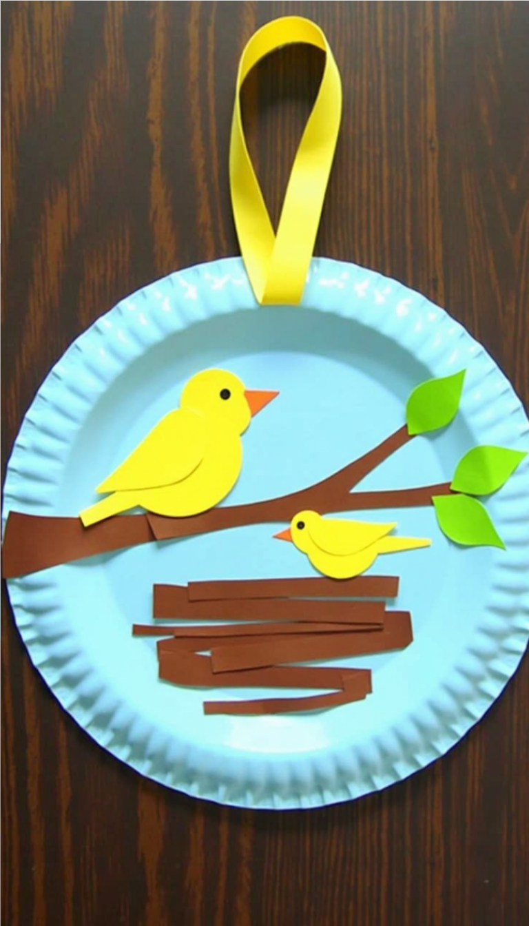 DIY spring craft for kids: a paper plate bird nest with two yellow paper birds on a brown branch, green leaves, and a yellow ribbon for hanging.