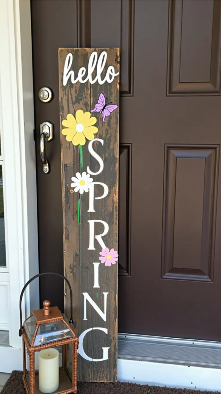 A rustic wooden sign with 'hello SPRING' in white letters, decorated with painted flowers and a butterfly, leaning against a brown front door.