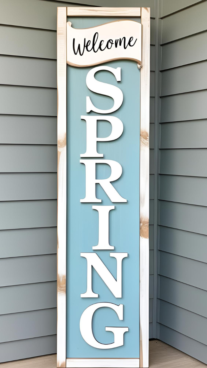 A tall wooden sign with a blue background and white frame, featuring 'Welcome SPRING' in raised white letters, leaning against a gray house exterior.
