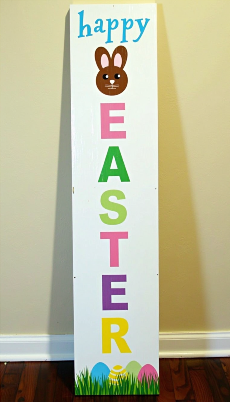 Vertical wooden Easter sign with a white background, featuring 'happy' in blue at the top, a brown bunny face in place of the letter 'A,' and colorful letters spelling 'EASTER.' The bottom is decorated with pastel Easter eggs nestled in green grass.