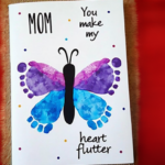 A handmade Mother's Day card featuring a butterfly created from a child's footprints in shades of blue, purple, and pink. The butterfly's body is painted black with curved antennae. Surrounding the butterfly are small, colorful dots. The card includes the words 'MOM' in bold letters on the left and the phrase 'You make my heart flutter' in playful, handwritten-style font.