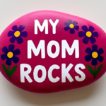 A smooth, oval-shaped rock painted bright pink with the words 'MY MOM ROCKS' in bold white letters. The design is decorated with four purple flowers with yellow centers and green stems, two on each side, adding a cheerful and decorative touch.