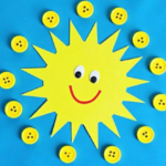 A cheerful sun craft made from yellow paper with a smiling face, surrounded by yellow buttons, all placed on a blue background. The buttons are arranged in a circular pattern around the sun, resembling sun rays.